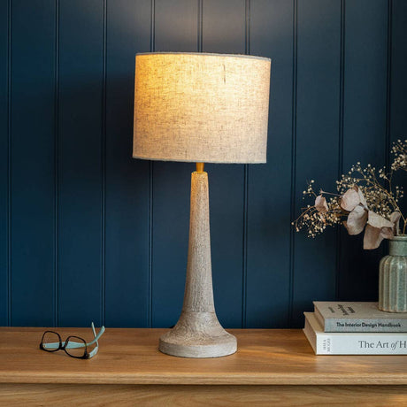 Table lamp with a textured base and beige shade on a wooden surface against a dark blue paneled wall.