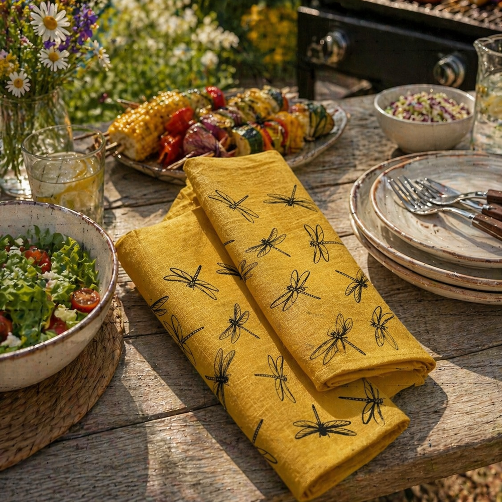 Outdoor dining setup with grilled food, salads, and a yellow Linen  Tea Towel with black dragonfly hand printed pattern.