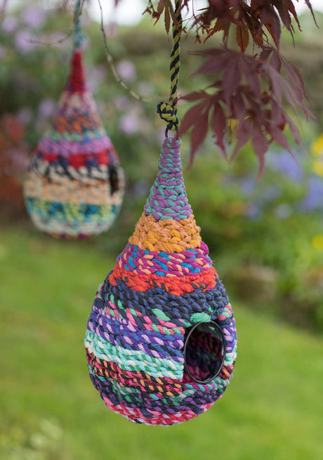Colorful woven birdhouse hanging from a tree branch with blurred greenery in the background.
