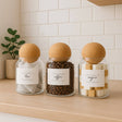 Three glass jars labeled 'tea', 'coffee', and 'sugar' on a wooden surface with a white tiled wall background.