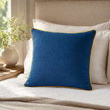 Plain deep blue linen cushion cover with mustard piping styled on a cream sofa.