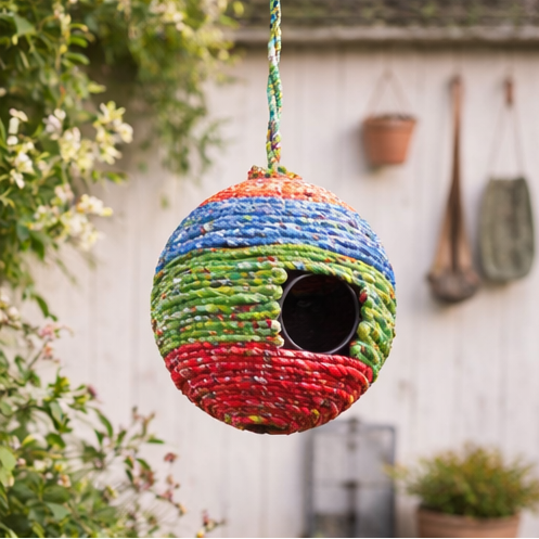 Colorful woven birdhouse hanging in a garden setting with flowers and plants.