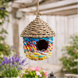 Colourful woven birdhouse hanging in a garden with flowers and trees.