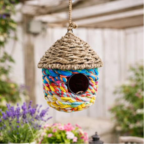 Colourful woven birdhouse hanging in a garden with flowers and trees.