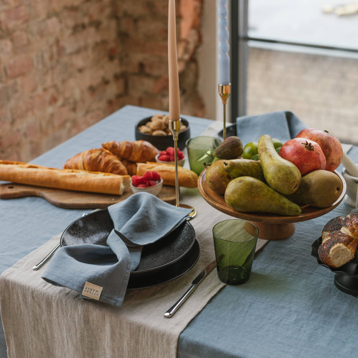 Relaxed, overhead table styling with mixed linen textures. Dining table set with croissants, pears, and other fruits on a blue tablecloth.