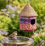 Colourful braided birdhouse with a thatched roof hanging above a birdbath in a garden setting.