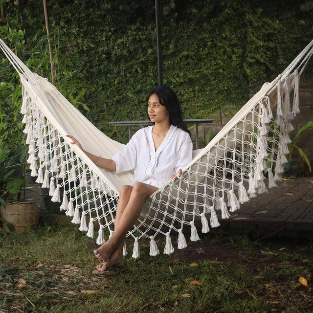 Woman sitting in a white hammock with tassels in a garden setting