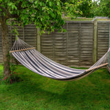 Striped hammock hanging between two trees in a garden with a wooden fence in the background.