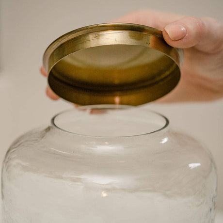 Hand holding a brass lid over a glass jar with a plain background