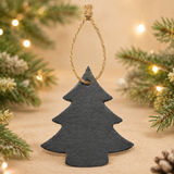 A dark gray slate Christmas tree tag hanging from a natural jute rope, set against a warm beige background with blurred pine branches and warm fairy lights.