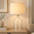 Table lamp with clear glass base and beige linen shade on a wooden surface.