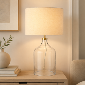 Table lamp with clear glass base and beige linen shade on a wooden surface.