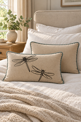 A linen cushion with black dragonfly print and a duck egg piping border, styled on a bed with other pillows and a textured blanket in a warm, neutral bedroom setting.