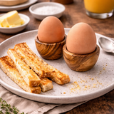 Two eggs in wooden holders with toast on a plate, accompanied by butter and a glass of orange juice.