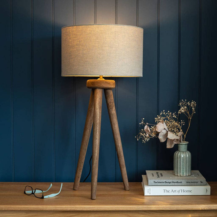 Wooden tripod lamp with beige shade on a wooden surface against a blue paneled wall.