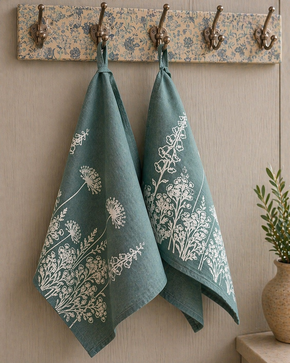 Two linen tea towels with white floral patterns hanging on a wooden hook rack against a beige wall.