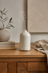 White ceramic diffuser on a wooden surface with a plant and books in the background