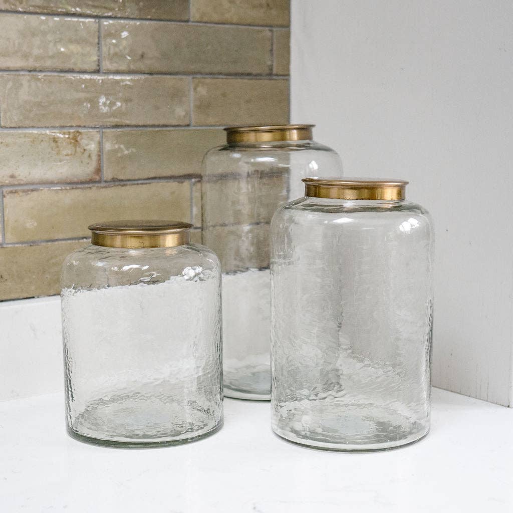 Three clear glass jars with gold lids on a white surface against a tiled wall.