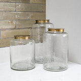 Three clear glass jars with gold lids on a white surface against a tiled wall.