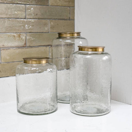 Three clear glass jars with gold lids on a white surface against a tiled wall.