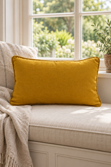 A mustard yellow rectangular linen pillow resting on a light beige upholstered window seat with a cream throw blanket, set in front of a bright window with green foliage visible outside.