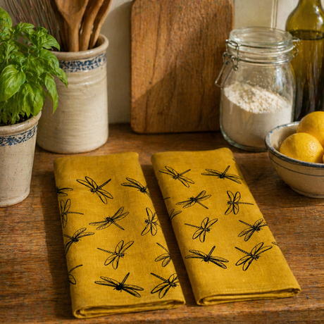 Yellow linen tea towels with black dragonfly patterns on a wooden kitchen counter with utensils and ingredients.