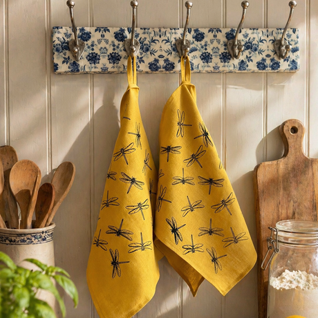Yellow linen tea towels with hand printed black dragonfly patterns hanging on a floral rack in a kitchen.