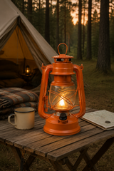 A glowing orange vintage-style lantern sits on a wooden camping table beside a metal mug and a notebook, with a tent and forest visible in the background at sunset.