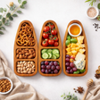 Three wooden snack platters with pretzels, fruits, vegetables, and cheese on a white surface.