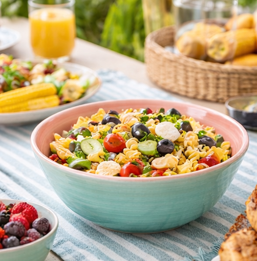 Colourful pasta salad in a teal bowl on a table with a basket of corn and a glass of orange juice.