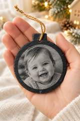 Personalised slate Christmas bauble with an engraved baby photograph, held in a hand to show its size.
