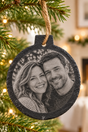 Personalised slate Christmas bauble with an engraved couple photograph, hanging from a fir branch by a jute loop.