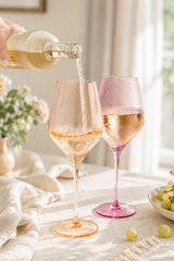 Person pouring white wine into two pink and peach wine glasses on a table with a soft, blurred background.