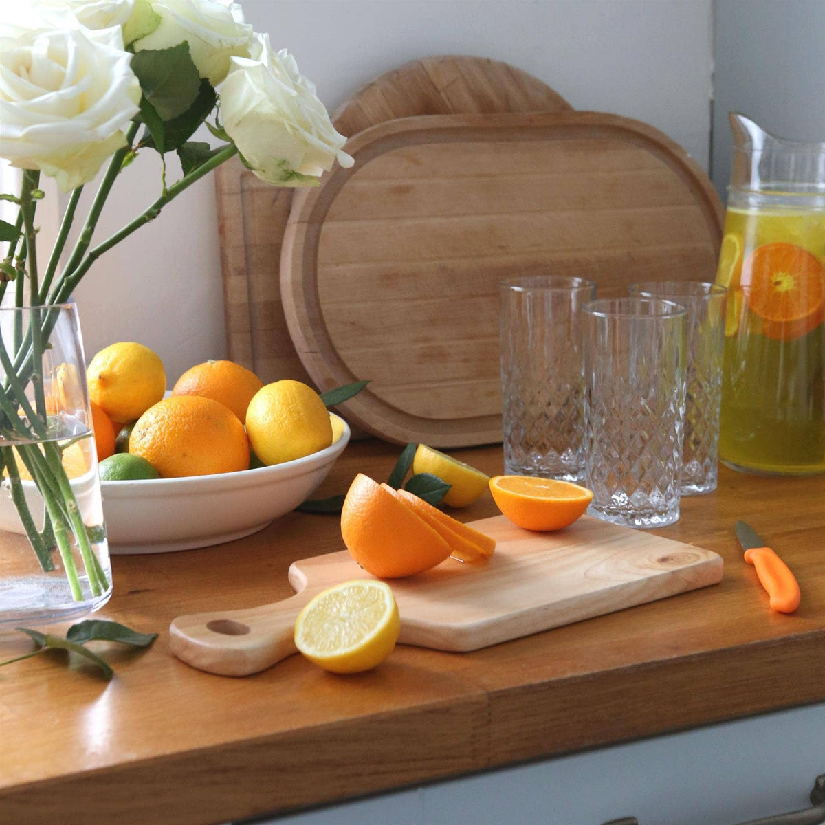 34 × 16cm rubberwood handled board with sliced orange and lemon on a kitchen counter beside glasses, a citrus drink jug and white roses.