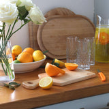 34 × 16cm rubberwood handled board with sliced orange and lemon on a kitchen counter beside glasses, a citrus drink jug and white roses.