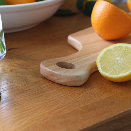 Close-up of the rounded wooden handle and hanging hole on the 34 × 16cm rubberwood board, beside a lemon half and sliced orange.