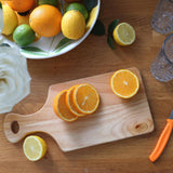 Overhead view of the 34 × 16cm rubberwood handled chopping board with sliced orange, beside lemon halves, a fruit bowl and an orange-handled knife.
