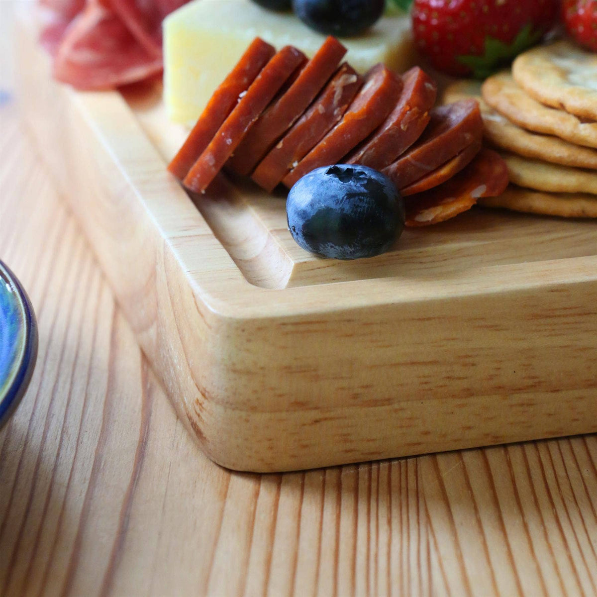 Close-up of the 30 × 20cm rubberwood board’s thick rounded edge and juice groove, with sliced sausage, cheese, crackers and berries.