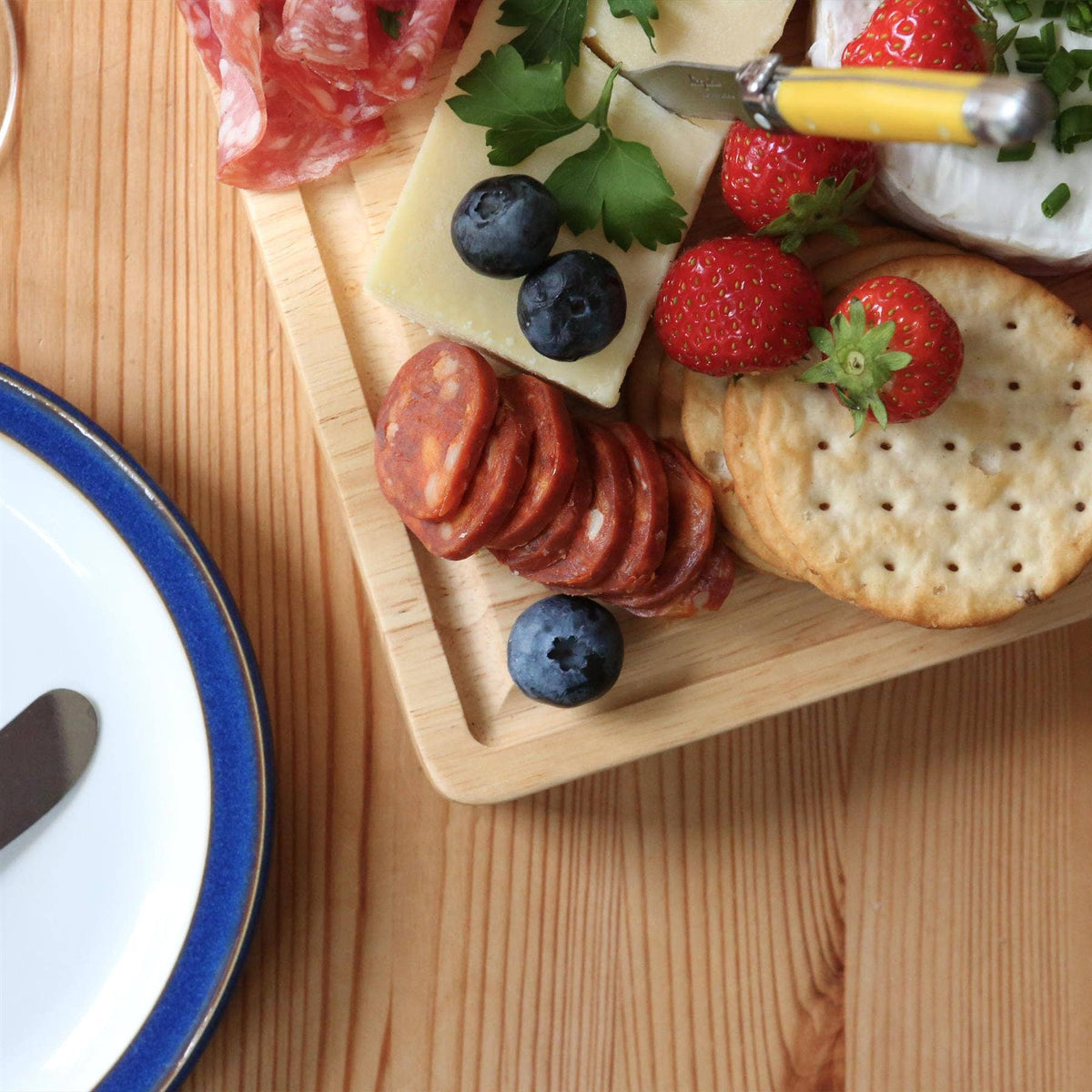 Overhead detail of the 30 × 20cm rubberwood board with cheese, sliced sausage, strawberries, blueberries and crackers beside a blue-rimmed plate.