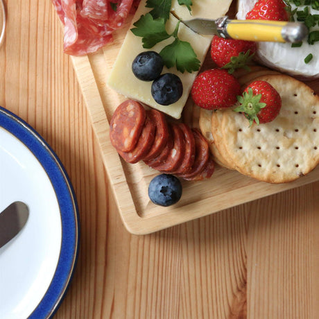 Overhead detail of the 30 × 20cm rubberwood board with cheese, sliced sausage, strawberries, blueberries and crackers beside a blue-rimmed plate.
