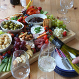 60 × 40cm rubberwood charcuterie board with cheeses, cured meats, crackers and fruit as white wine is poured beside it.