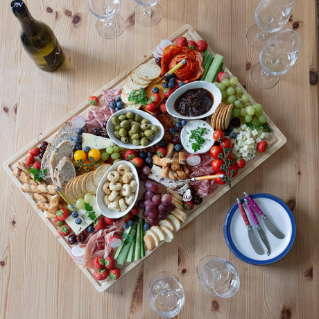 Overhead view of a 60 × 40cm rubberwood grazing board filled with cheese, cured meats, fruit, crackers, olives and dips.