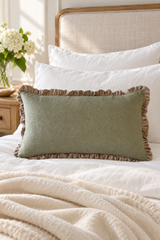 A sage green linen lumbar pillow with a brown ruffled border, styled on a bed with white pillows and a cream blanket in a softly lit bedroom.