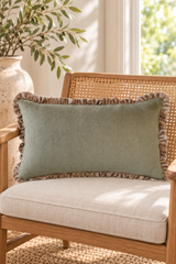 A sage green lumbar linen pillow with a brown ruffled trim, styled on a light beige upholstered wooden armchair in a bright, sunlit room.