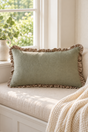 A rectangular sage green linen pillow with ruffled trim rests on a cream-colored window seat cushion, with a cream knit blanket draped over the side and a bright window in the background.