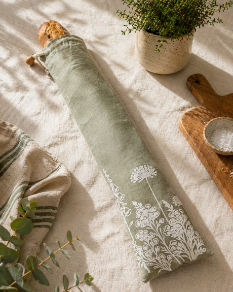 sage Green linen bread bag with white floral patterns on a light wooden surface with a plant and wooden cutting board.