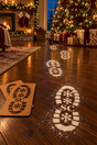 Snowy Santa boot footprints leading towards Christmas presents and a decorated tree, with the wooden stencil pair beside them.