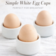Three brown eggs in white ceramic cups on a white textured surface with text 'Simple White Egg Cups Perfect for Breakfast'.