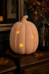 A decorative carved terracotta pumpkin with star-shaped cutouts glowing warm yellow from inside, displayed on a dark wooden surface with autumn leaves and a dark glass vase in the background.