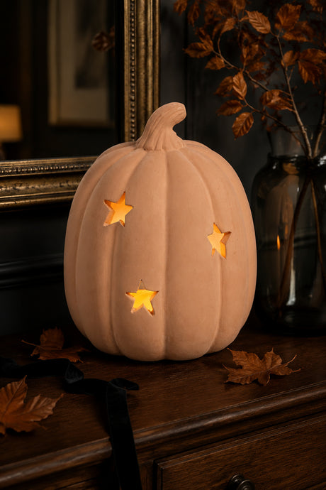 A decorative carved terracotta pumpkin with star-shaped cutouts glowing warm yellow from inside, displayed on a dark wooden surface with autumn leaves and a dark glass vase in the background.